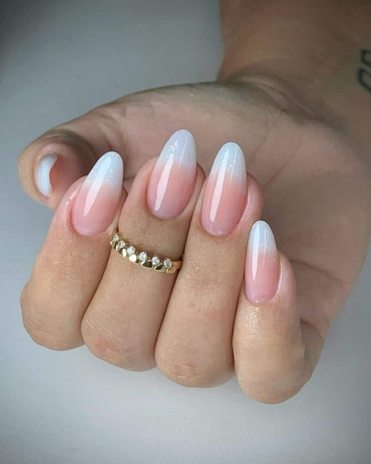 Mão com unhas longas no formato amendoado, exibindo a manicure baby-boomer com efeito degradê suave do rosa (leitoso) para o branco nas pontas, com um anel dourado.