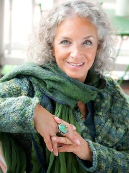 Mulher sorridente com cabelos grisalhos e cacheados, usando casaco e cachecol em tons de verde esmeralda. A cor verde confere frescor e modernidade ao visual grisalho em 2025.