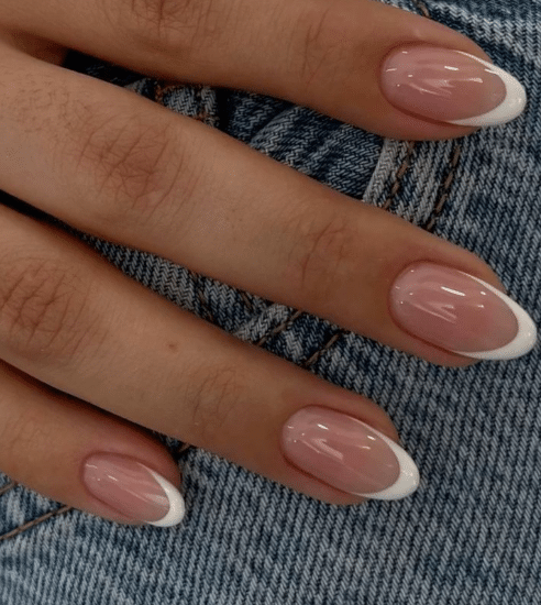 Mãos com unhas francesinhas clássicas em esmalte nude e pontas brancas sobre tecido jeans azul.