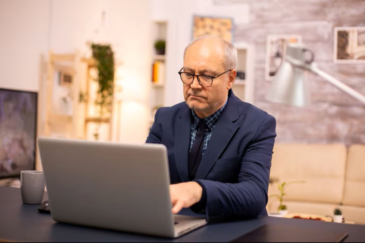 Homem sênior concentrado trabalhando em laptop em escritório moderno, representando profissões para maiores de 60 anos.