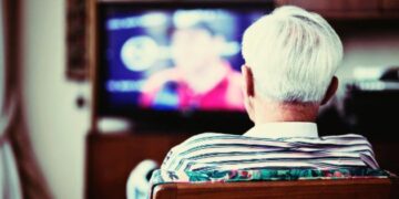 Pessoa idosa assistindo TV de costas, sentada em um sofá.