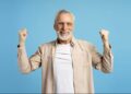 Idoso sorridente, com barba branca e óculos, celebrando com os punhos erguidos, em fundo azul.