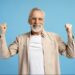Idoso sorridente, com barba branca e óculos, celebrando com os punhos erguidos, em fundo azul.