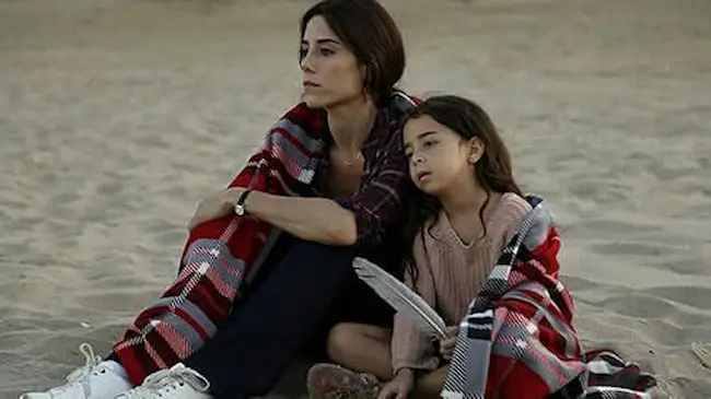 Mãe e filha sentadas na areia, compartilhando um momento de ternura na novela turca “Mãe”.