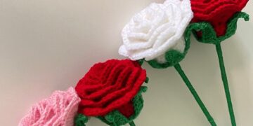 Rosa de crochê enrolada nas cores vermelho, branco e rosa, dispostas em diagonal, com foco no detalhe dos pontos e haste verde. Tutorial de artesanato fácil.