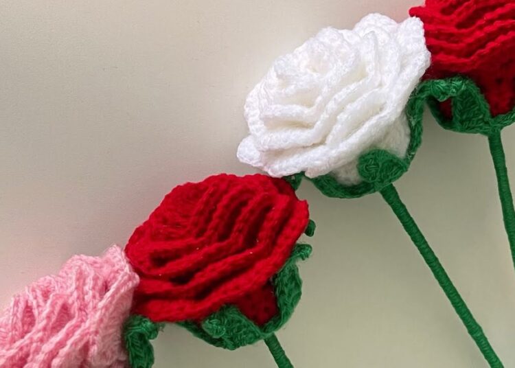 Rosa de crochê enrolada nas cores vermelho, branco e rosa, dispostas em diagonal, com foco no detalhe dos pontos e haste verde. Tutorial de artesanato fácil.