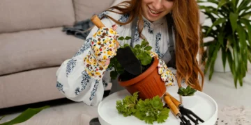 Mulher sorridente cuidando de uma planta em vaso dentro de casa, usando luvas floridas e ferramentas de jardinagem.