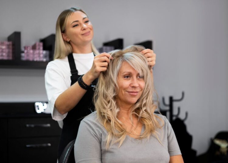 Cabeleireira loira em salão de beleza finalizando o penteado de uma cliente madura com cabelos claros, realçando um corte que afina o rosto.