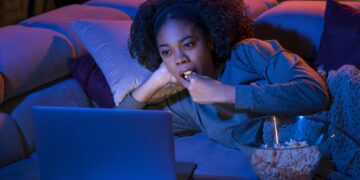 Mulher deitada no sofá com laptop e comendo pipoca à noite