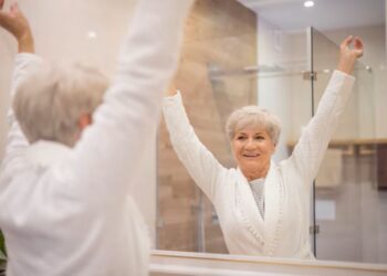 Imagem de uma mulher idosa sorrindo e levantando os braços diante do espelho, refletindo sobre autocuidado e bem-estar.