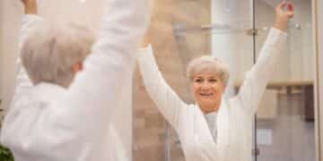 Imagem de uma mulher idosa sorrindo e levantando os braços diante do espelho, refletindo sobre autocuidado e bem-estar.