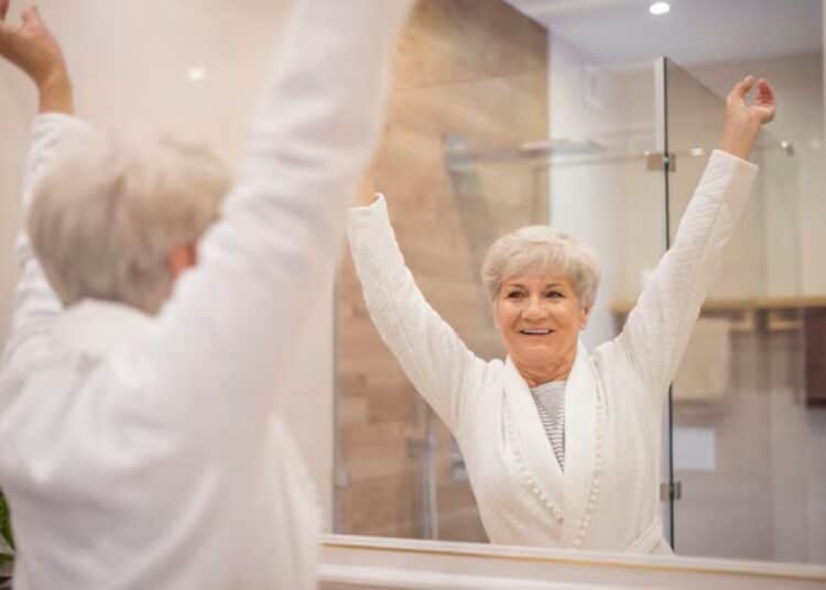 Imagem de uma mulher idosa sorrindo e levantando os braços diante do espelho, refletindo sobre autocuidado e bem-estar.