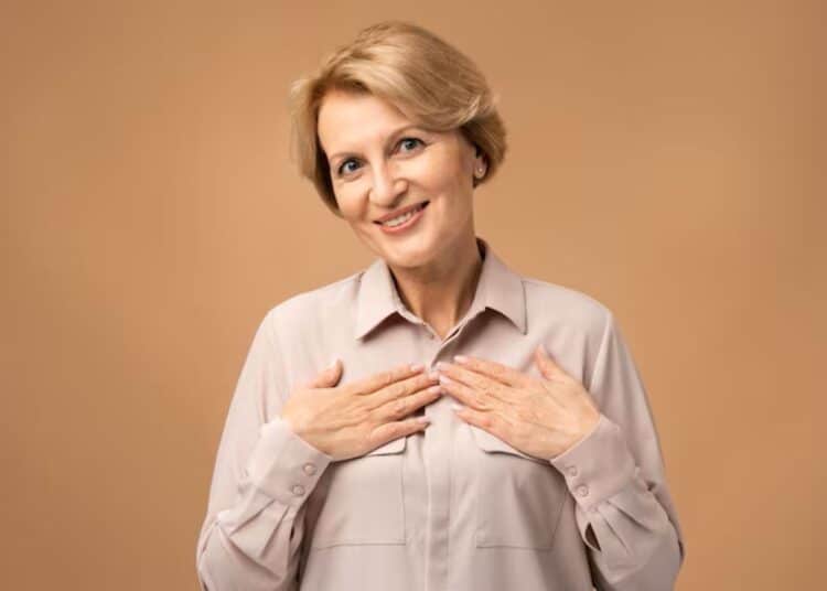 Mulher sorridente de cabelos loiros curtos, com mais de 50 anos, usando uma blusa em tom neutro e as mãos no peito, irradiando autoconfiança e a beleza da maturidade.