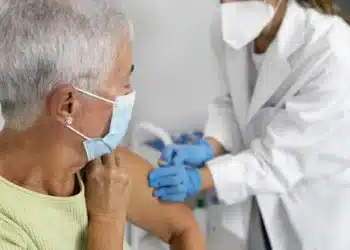 Idosa recebendo uma vacina de um profissional de saúde, com ambos usando máscara de proteção