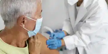 Idosa recebendo uma vacina de um profissional de saúde, com ambos usando máscara de proteção