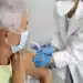 Idosa recebendo uma vacina de um profissional de saúde, com ambos usando máscara de proteção