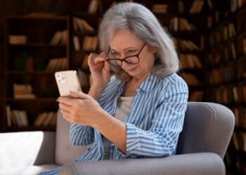 Mulher idosa sorrindo, mexendo no celular em casa, com estantes de livros ao fundo. A imagem sugere a busca por mensagens e orações para postar no status de fim de semana.