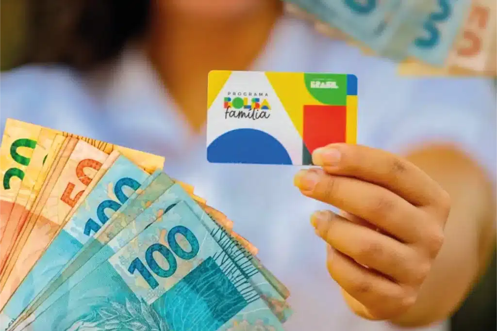 Bolsa Família 2026 Pessoa segurando o cartão do Programa Bolsa Família com notas de dinheiro ao fundo, representando a consulta do pagamento de janeiro de 2026.