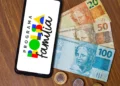 Celular exibindo logo do Programa Bolsa Família ao lado de cédulas e moedas de real sobre mesa de madeira.