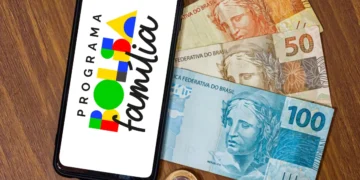 Celular exibindo logo do Programa Bolsa Família ao lado de cédulas e moedas de real sobre mesa de madeira.