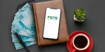 Smartphone mostrando logo do FGTS sobre caderno, dinheiro, caneta, xícara de café e planta