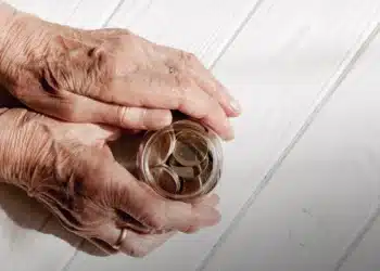 Mãos envelhecidas de um idoso segurando um pote transparente cheio de moedas em superfície branca de madeira