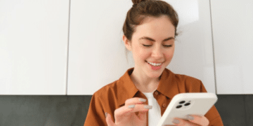 Mulher sorridente com cabelo preso usando smartphone branco em ambiente interno com armários brancos ao fundo