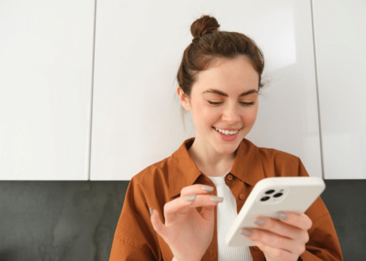Mulher sorridente com cabelo preso usando smartphone branco em ambiente interno com armários brancos ao fundo