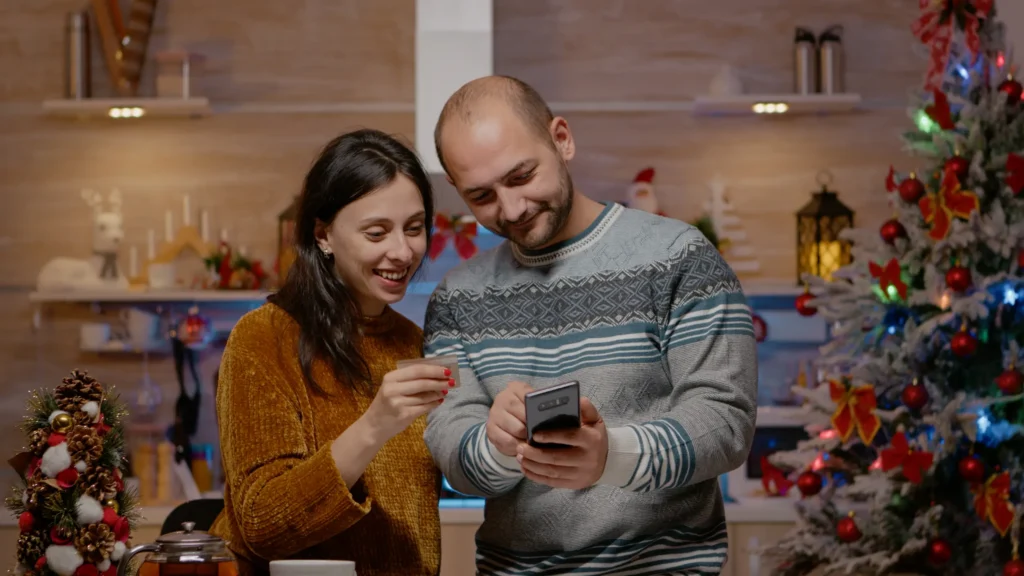 Mensagens de Natal Casal sorrindo e compartilhando um momento de alegria, enquanto visualizam algo em um smartphone, com decoração de Natal ao fundo.