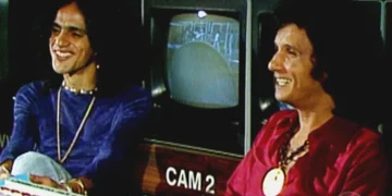 Caetano Veloso e Roberto Carlos sorrindo juntos durante uma gravação de TV, com uma tela de monitor ao fundo, simbolizando a amizade entre eles.