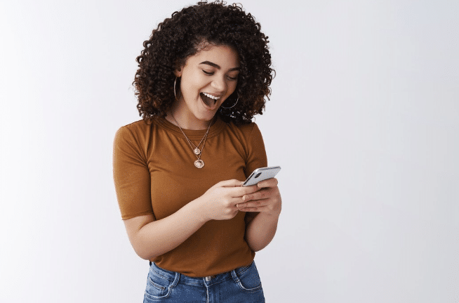 Mulher sorrindo, com cabelo cacheado e camiseta marrom, olhando para celular na mão