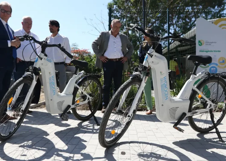 Bicicletas elétricas brancas movidas a hidrogênio expostas com pessoas observando ao fundo