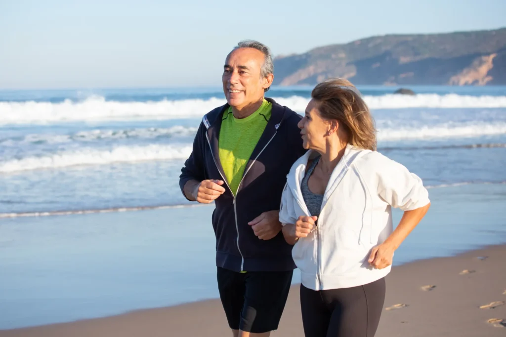 Exercícios para idosos no verão Casal de idosos caminhando na praia durante o verão, praticando exercício físico ao ar livre.