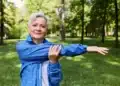 Idosa praticando exercícios ao ar livre, em um ambiente natural, usando um casaco azul, realizando alongamento de braço.