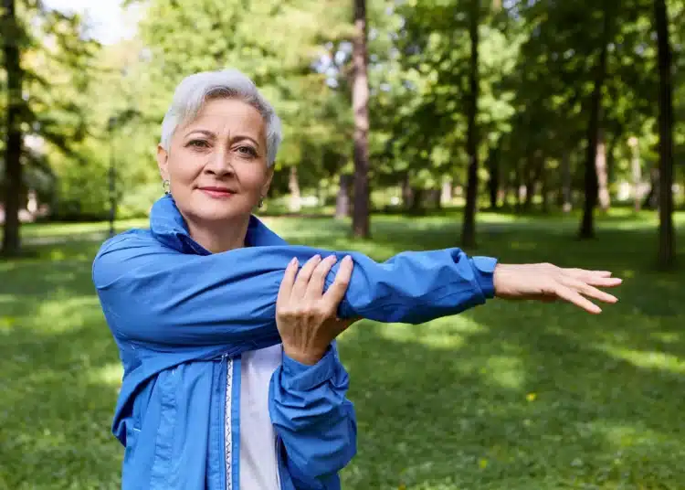 Idosa praticando exercícios ao ar livre, em um ambiente natural, usando um casaco azul, realizando alongamento de braço.