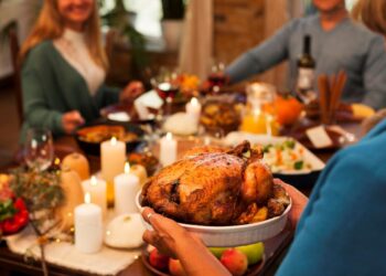 Close-up das mãos de uma pessoa segurando um travessa branca com um frango assado dourado e suculento, em uma mesa de Natal decorada com velas acesas, frutas e convidados sorridentes ao fundo.
