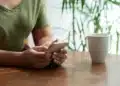 Pessoa segurando um smartphone com uma xícara de café sobre a mesa, rodeada por plantas verdes em um ambiente acolhedor.