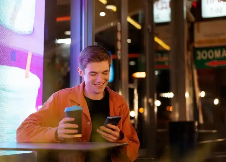 Jovem sorrindo à noite enquanto segura um café e lê mensagem no celular, representando frases de boa noite para WhatsApp.
