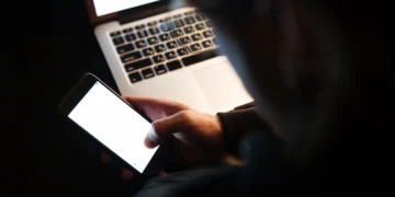 Pessoa usando celular à noite com laptop ao fundo, simbolizando o risco de fraudes online durante as festas de fim de ano.