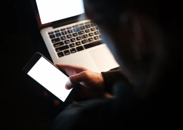 Pessoa usando celular à noite com laptop ao fundo, simbolizando o risco de fraudes online durante as festas de fim de ano.