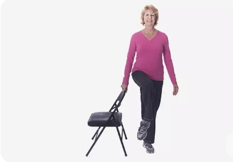 Mulher idosa com blusa rosa fazendo exercício de equilíbrio segurando uma cadeira preta