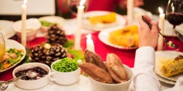 Mesa de jantar festiva com toalha vermelha, velas acesas, pratos com milho, ervilhas e batatas. Uma pessoa de camisa branca aparece em primeiro plano servindo-se.