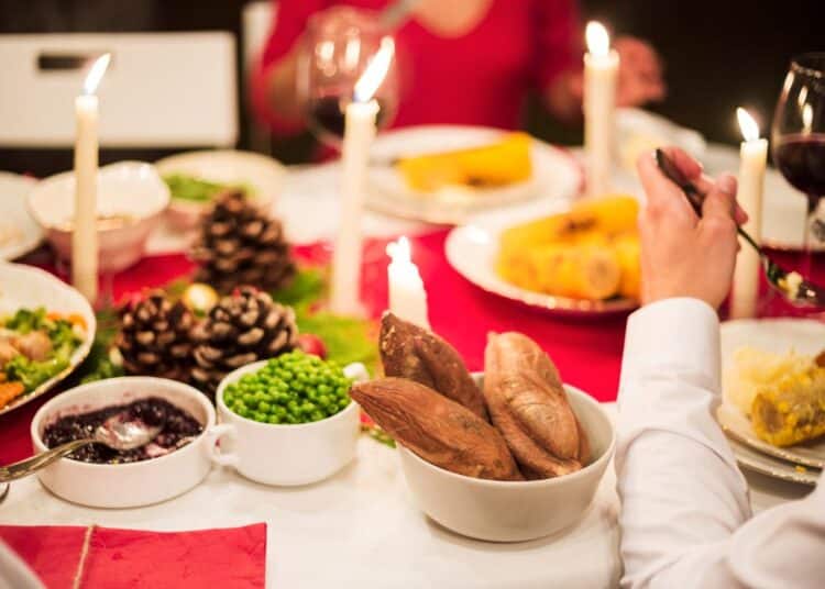 Mesa de jantar festiva com toalha vermelha, velas acesas, pratos com milho, ervilhas e batatas. Uma pessoa de camisa branca aparece em primeiro plano servindo-se.