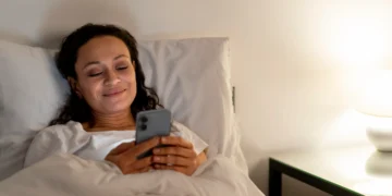 Mulher sorrindo na cama enquanto envia uma mensagem no celular, preparando-se para descansar após um dia cansativo de segunda-feira.