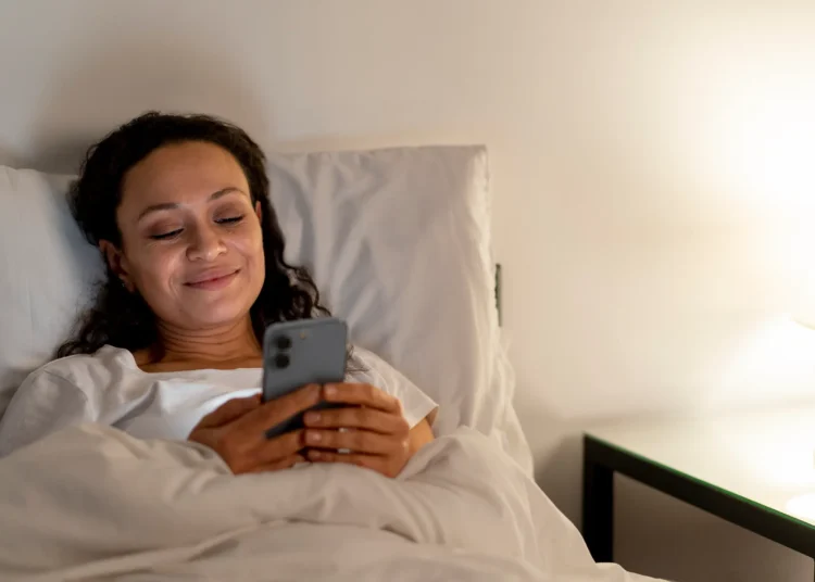Mulher sorrindo na cama enquanto envia uma mensagem no celular, preparando-se para descansar apĂ³s um dia cansativo de segunda-feira.