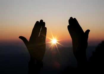 Silhueta de mãos elevadas em frente ao pôr do sol, transmitindo um momento de conexão espiritual e fé.