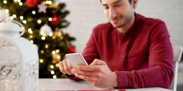 Homem usando smartphone em ambiente natalino com árvores de Natal e luzes brilhantes, perfeito para mensagens de fim de ano.