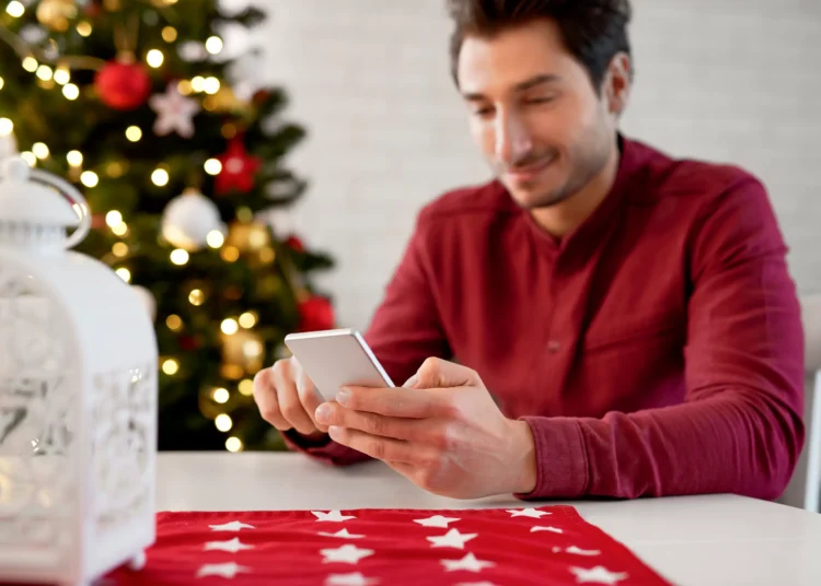 Homem usando smartphone em ambiente natalino com árvores de Natal e luzes brilhantes, perfeito para mensagens de fim de ano.