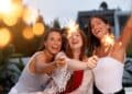 Três amigas sorridentes celebrando ao ar livre durante o entardecer, segurando velas faíscas (sparklers) acesas que criam pontos de luz brilhantes.