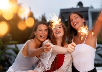 Três amigas sorridentes celebrando ao ar livre durante o entardecer, segurando velas faíscas (sparklers) acesas que criam pontos de luz brilhantes.